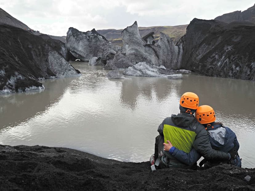 A couple in helmets sitting with arms around each other looking out at a glacier and its lagoon