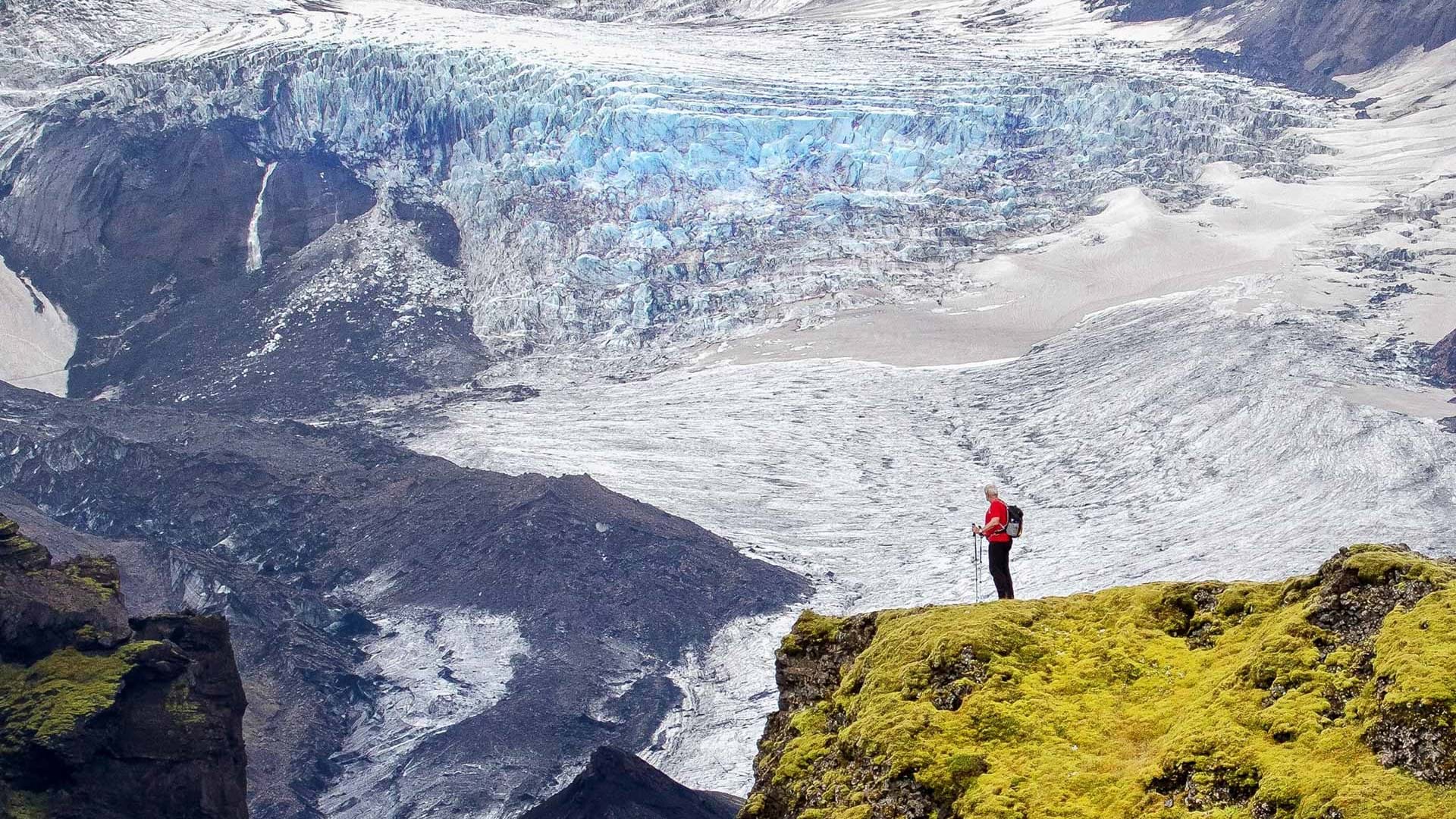 A man standing on a moss covered cliff looking at a magnificent glacier in the background