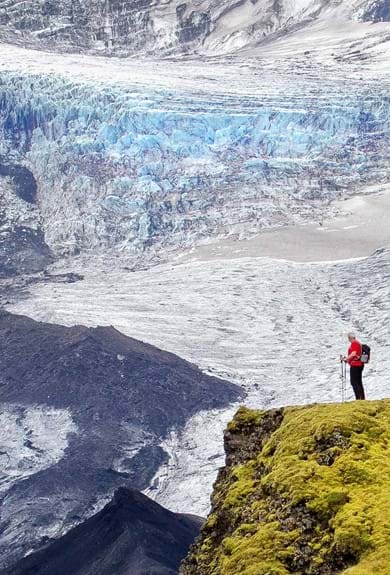 A man standing on a moss covered cliff looking at a magnificent glacier in the background