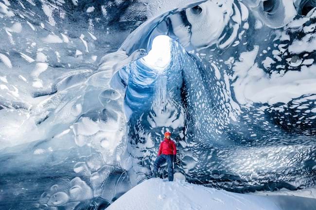A person in a bright red jacket stands at the entrance of a stunning ice cave, with the natural blue hues of the glacial ice swirling around them, creating an ethereal and captivating scene.