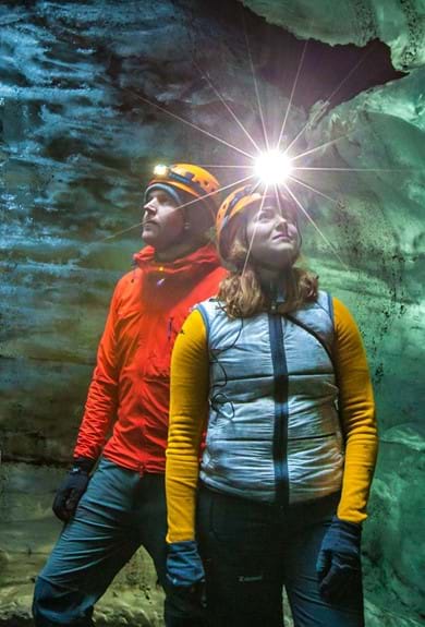A couple standing in an ice cave in Iceland, wearing helmets with headlamps and gazing upwards