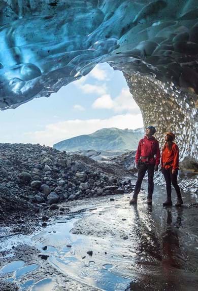Glacier Ice Cave in Skaftafell, two persons dressed in red standing in an ice cave surrounded by crystal blue ice with the cave opening in the background.