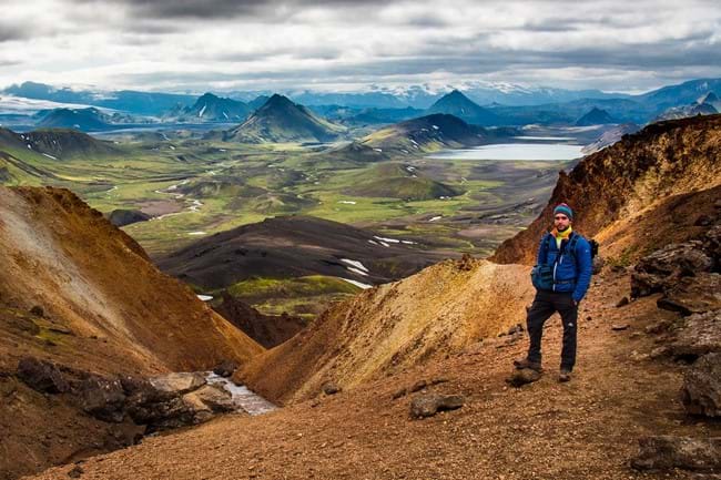 A man standing on a yellow mountain posing with the Icelandic nature and blue mountains in the far back.